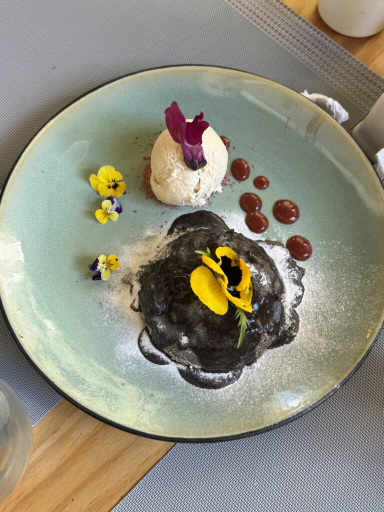 A dessert is elegantly presented on a light green plate. It features a dark chocolate lava cake topped with a yellow edible flower and dusted with powdered sugar. Beside it is a scoop of vanilla ice cream garnished with a purple flower. There are small dollops of red sauce on the plate, along with additional small yellow and purple flowers for decoration. The plate is set on a wooden table with a gray placemat.