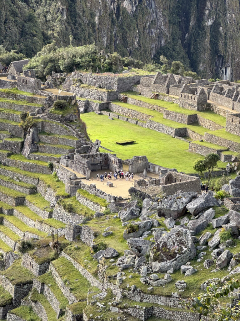 The image shows the ancient Incan city of Machu Picchu, located in the Andes Mountains of Peru. The scene includes terraced fields, stone structures, and ruins set against a backdrop of steep, lush green mountains. A group of people is visible exploring the site, highlighting its historical and archaeological significance.