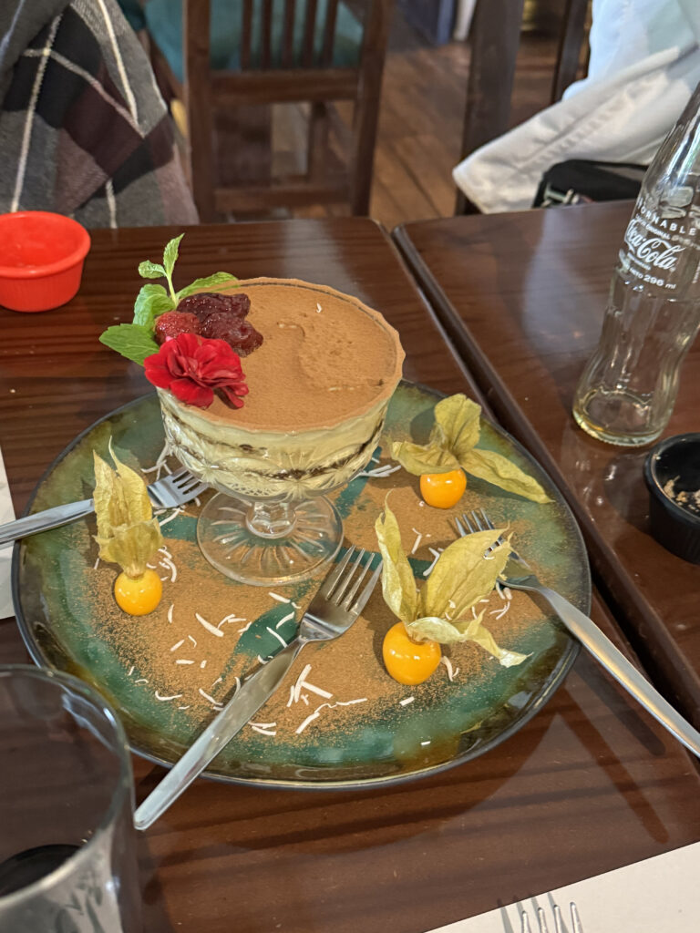A dessert is elegantly presented on a green and brown plate. The dessert, possibly tiramisu, is in a glass dish and topped with a red flower, mint leaves, and a dollop of jam. The plate is decorated with three physalis fruits and sprinkled with white shavings. Three forks are placed around the dessert. A glass bottle and a red container are visible in the background.