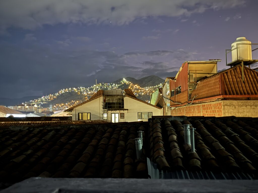 A nighttime cityscape featuring rooftops with red tiles in the foreground. In the background, a hillside is illuminated with numerous small lights, creating a sparkling effect against the night sky. The sky is partly cloudy, adding depth to the scene.