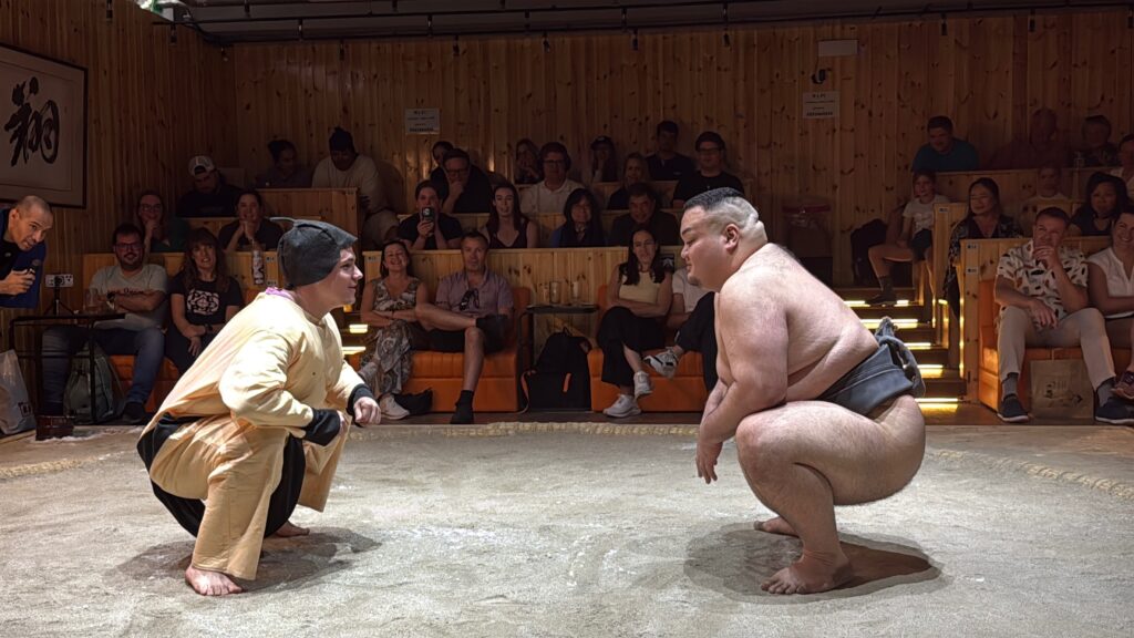 A sumo wrestler and a person in a padded suit are squatting and facing each other in a sumo ring. The setting is indoors with a wooden interior, and there is an audience seated around the ring, watching the match.