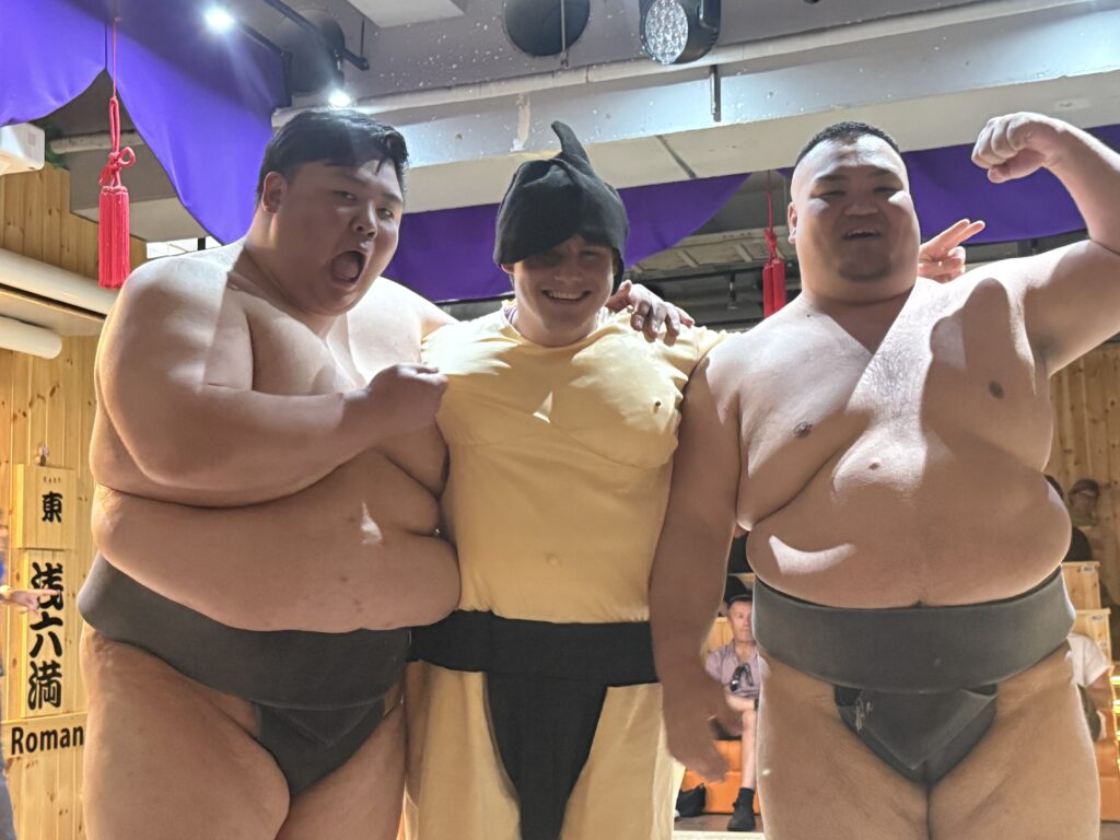 Three sumo wrestlers are posing together in a brightly lit room. Two of them are wearing traditional sumo attire, while the person in the middle is dressed in a yellow costume with a black hat. They are smiling and appear to be in a friendly, celebratory mood. The background features wooden paneling and purple drapery.