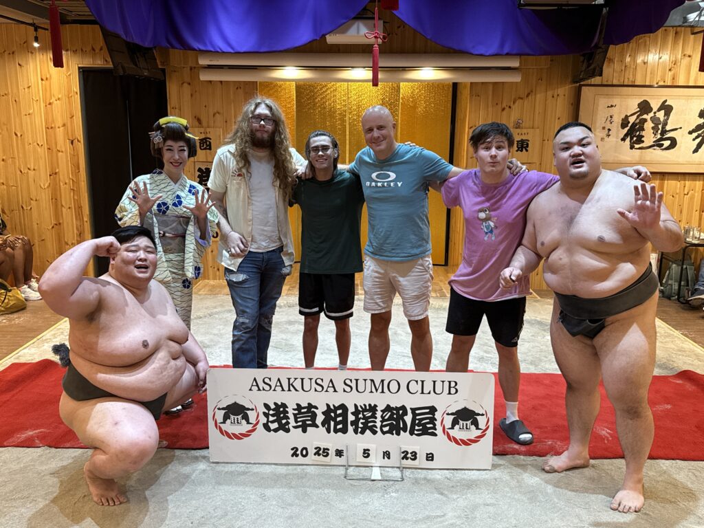 A group of people is posing together at the Asakusa Sumo Club. Two sumo wrestlers in traditional attire are on either side of the group. In the center, there are four people, including a person in a kimono and three casually dressed individuals. They are all standing on a sandy surface with a sign in front of them that reads "Asakusa Sumo Club" in English and Japanese. The background features wooden walls and traditional Japanese decor.