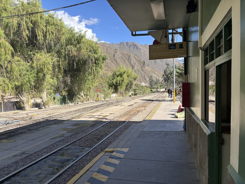 A small train station platform with tracks running alongside it. There is a sign for a VIP lounge above the platform. The area is surrounded by lush green trees and mountains in the background. A few people are visible in the distance on the platform. The sky is clear and blue.