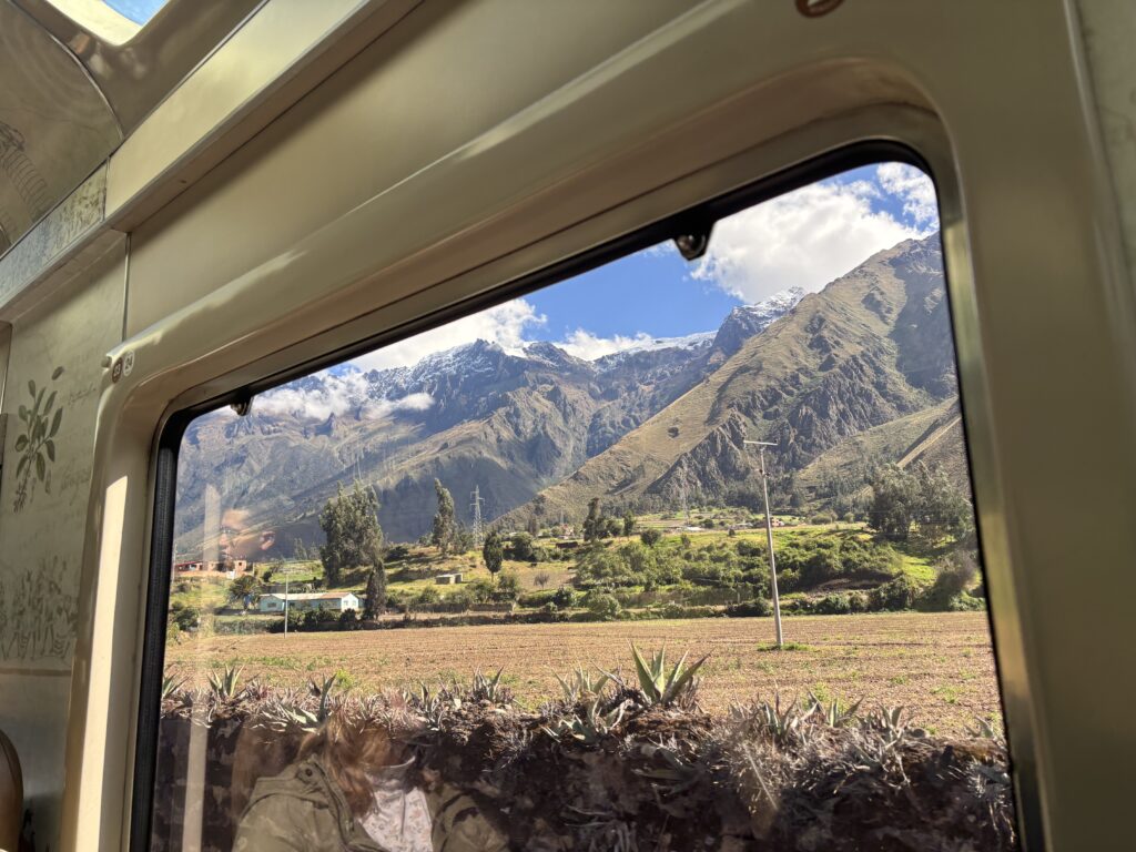 The image shows a scenic view of a mountainous landscape seen through a train window. The mountains are partially covered with snow and surrounded by clouds. In the foreground, there are fields and a few small buildings, with some trees scattered across the landscape. The interior of the train is visible, including part of the window frame and reflections on the glass.