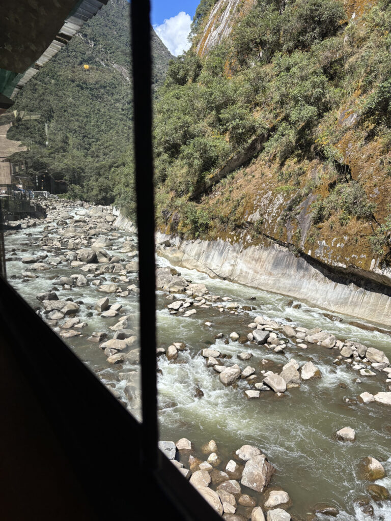View from a window showing a rocky river flowing through a mountainous landscape. The river is surrounded by large rocks and bordered by steep, tree-covered hills. The sky is clear with a few clouds visible in the background.