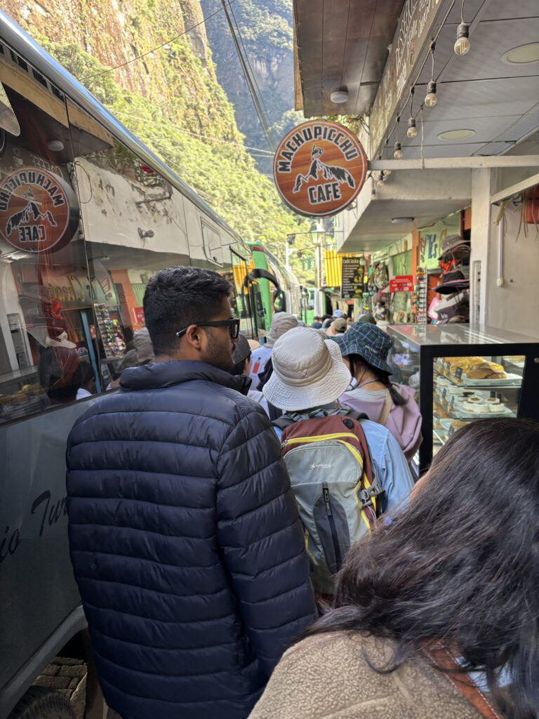 The image shows a crowded street scene with people walking near a bus. A sign for "Machu Picchu Cafe" is visible above the crowd. The area is surrounded by tall, green-covered cliffs, suggesting a mountainous location. Some people are wearing hats and backpacks, indicating they might be tourists. There is a display case with pastries on the right side.