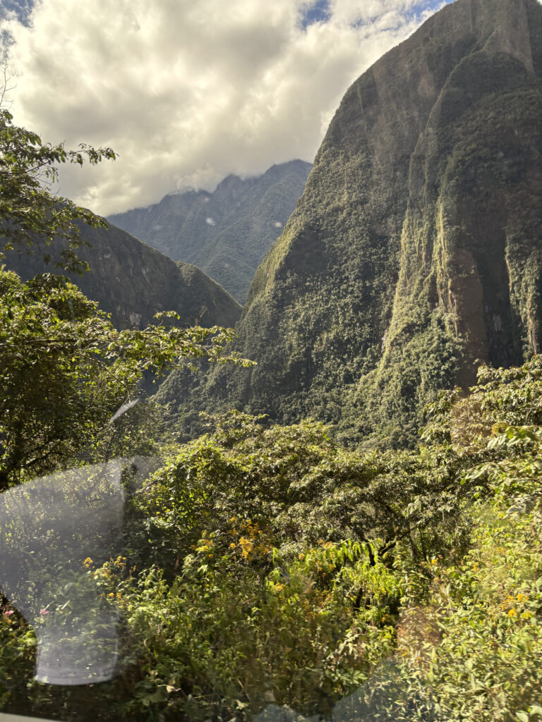 A lush, green mountainous landscape with steep, forested slopes under a partly cloudy sky. The foreground features dense vegetation, including trees and shrubs, with sunlight filtering through the clouds, casting light and shadows on the scene.