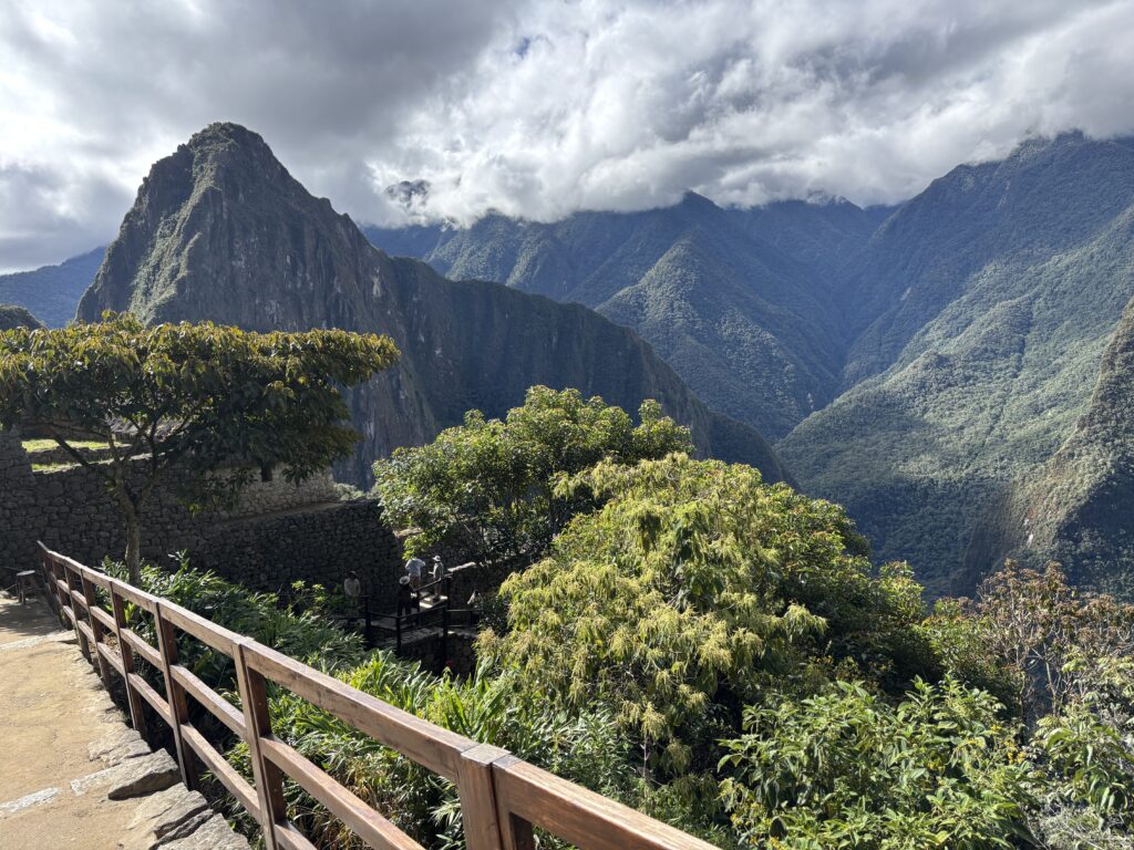 A scenic view of a mountainous landscape with lush green vegetation in the foreground. A wooden railing lines a stone pathway on the left, leading towards a stone structure partially visible among the trees. In the background, a prominent mountain peak rises under a cloudy sky, with other mountains extending into the distance.