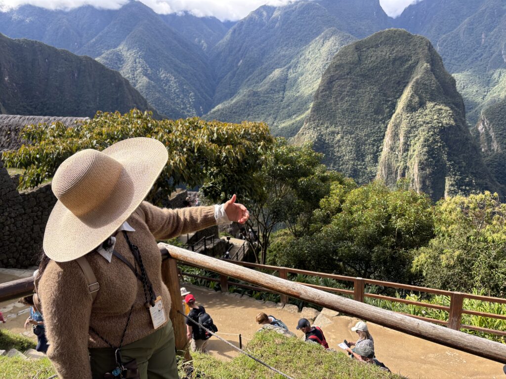 A person wearing a large straw hat and a brown sweater is gesturing towards a scenic view of lush green mountains. The person is standing near a wooden railing, and there are other people sitting on the ground, possibly tourists, with some holding maps. The background features a dramatic landscape of steep, forested mountains under a partly cloudy sky.