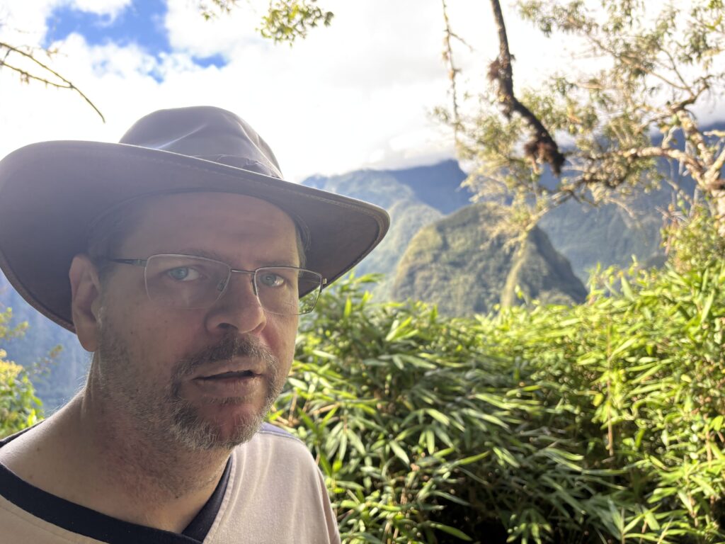 A man wearing a wide-brimmed hat and glasses is standing outdoors in a lush, green environment. Behind him, there are dense green plants and a mountainous landscape under a partly cloudy sky.