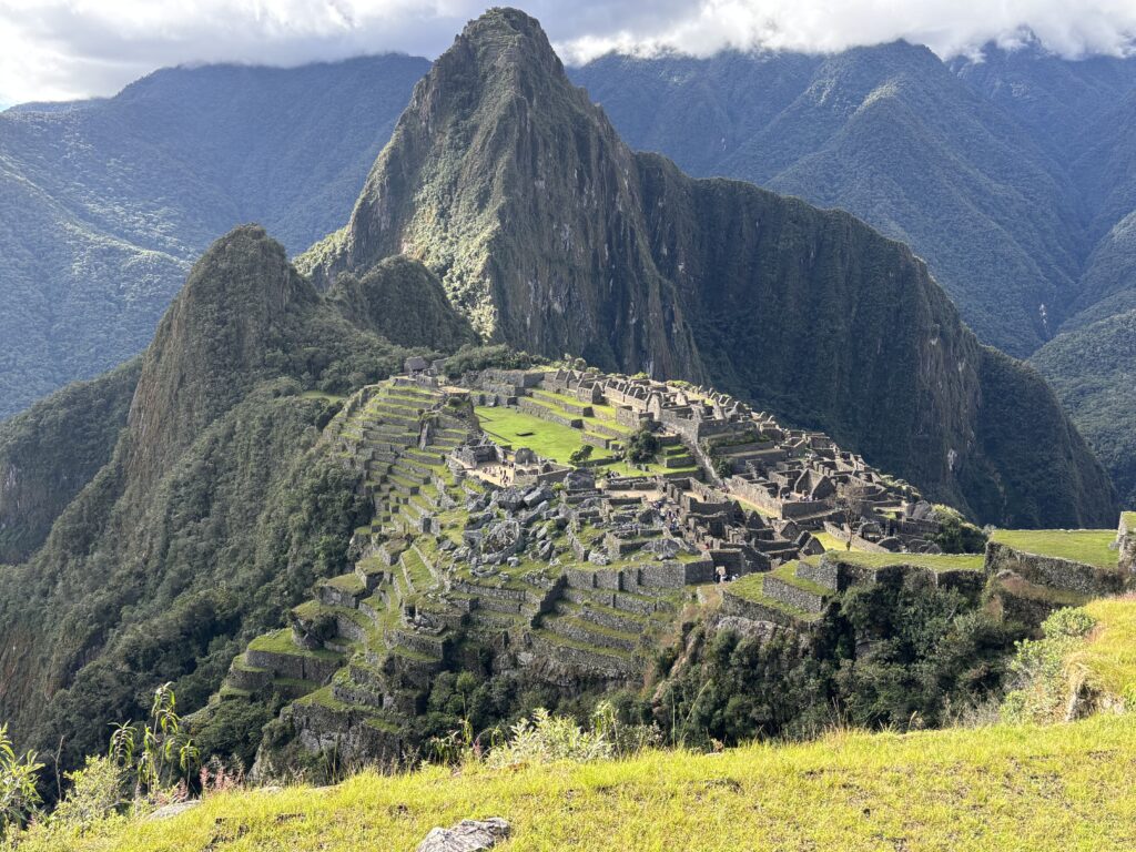 The image shows the ancient Incan city of Machu Picchu, located in the Andes Mountains of Peru. The ruins are set on a mountain ridge with terraced fields and stone structures. In the background, steep, lush green mountains rise under a partly cloudy sky.
