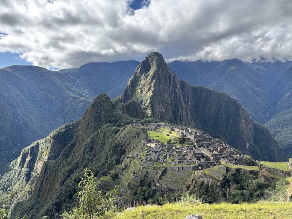The image shows the ancient Incan city of Machu Picchu, situated on a mountain ridge with lush green terraces and stone structures. The background features towering mountains under a partly cloudy sky.