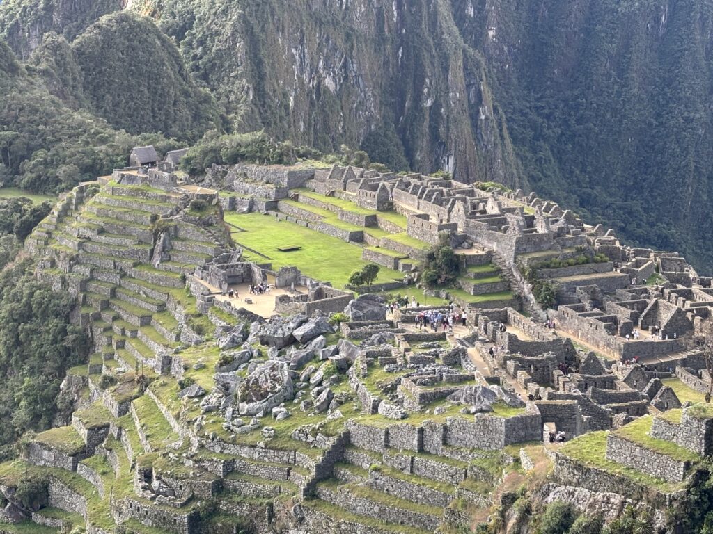 Aerial view of Machu Picchu, an ancient Incan city located in the Andes Mountains of Peru. The image shows the stone structures and terraces built into the mountainside, surrounded by lush green vegetation and steep cliffs. Visitors can be seen exploring the site.