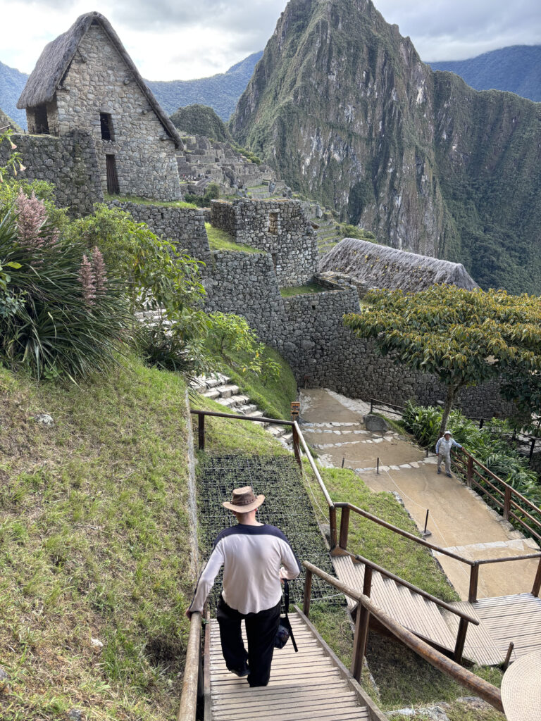 A person wearing a hat is walking down a wooden staircase at Machu Picchu, an ancient Incan city in Peru. The scene includes stone structures with thatched roofs, surrounded by lush green mountains and vegetation. The sky is cloudy, adding to the dramatic landscape.