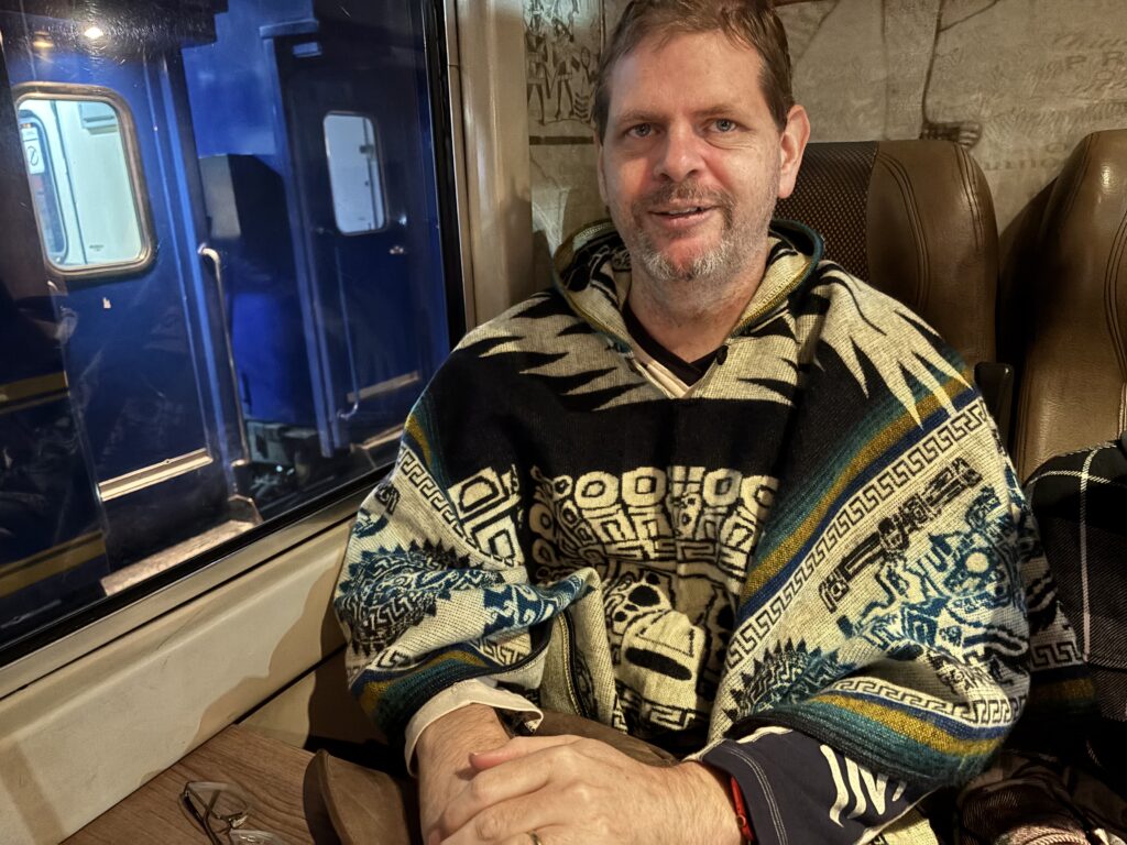 A person is sitting inside a train, wearing a colorful patterned poncho. The train's interior is visible, with brown seats and a window showing the exterior of another blue train car. The person is smiling and has their hands resting on a table. Glasses are placed on the table.