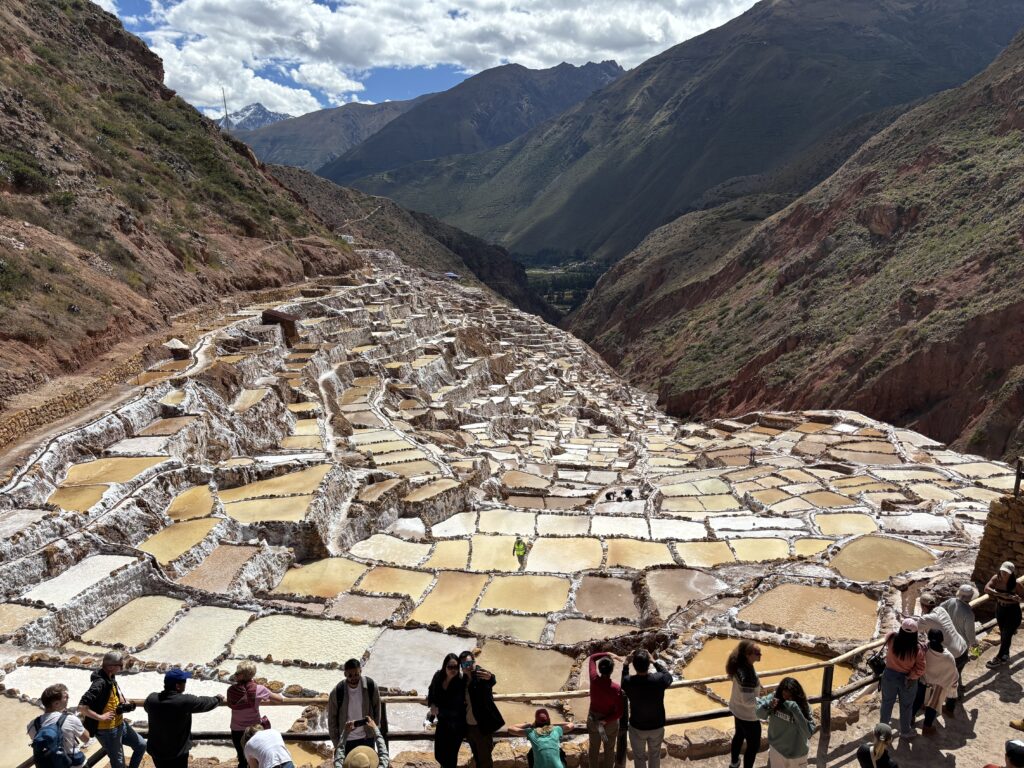 The image shows a vast landscape of terraced salt evaporation ponds, known as salt pans, situated on a hillside. The pans are filled with water in various shades of white and brown, creating a patchwork effect. In the foreground, a group of people are standing along a wooden railing, observing the scene. The background features rugged mountains under a partly cloudy sky.