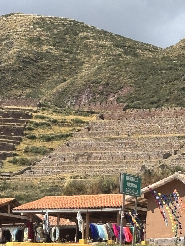 The image shows a hillside with terraced stone structures, likely agricultural terraces. In the foreground, there is a market stall with colorful textiles and crafts on display. A sign in Spanish reads "REDUCE REUSA RECICLA," which translates to "Reduce, Reuse, Recycle." The sky is overcast, and the landscape is covered with patches of grass and vegetation.