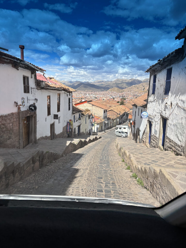 A narrow cobblestone street descends between whitewashed buildings with wooden balconies and tiled roofs. The street is lined with stone walls and leads to a view of a cityscape nestled in a valley surrounded by mountains. The sky is bright blue with scattered clouds.