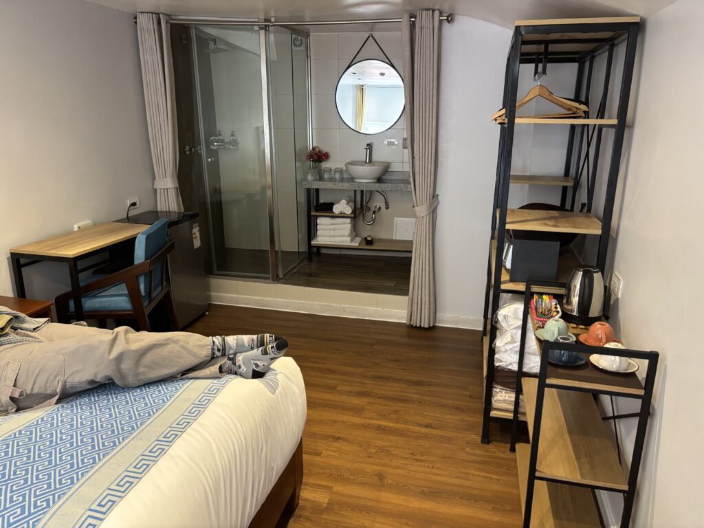 The image shows a hotel room with a modern design. In the foreground, there is a bed with a blue and white patterned runner. A person is lying on the bed, with their legs visible, wearing light-colored pants and patterned socks. To the left, there is a small desk with a blue chair and a mini-fridge underneath. In the background, there is a glass-enclosed shower and a sink with a round mirror above it. On the right, there is a shelving unit with hangers, a kettle, cups, and other amenities. The room has wooden flooring and light-colored walls.