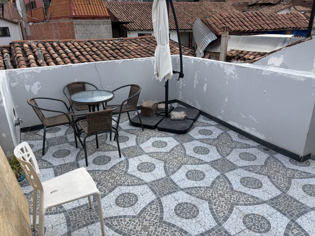 A rooftop terrace with a patterned tile floor featuring a circular design. There is a round glass-top table with four wicker chairs around it. An umbrella is closed and secured with stones on a stand. A white plastic chair is also present. The terrace is surrounded by a low wall, and in the background, there are buildings with red-tiled roofs.
