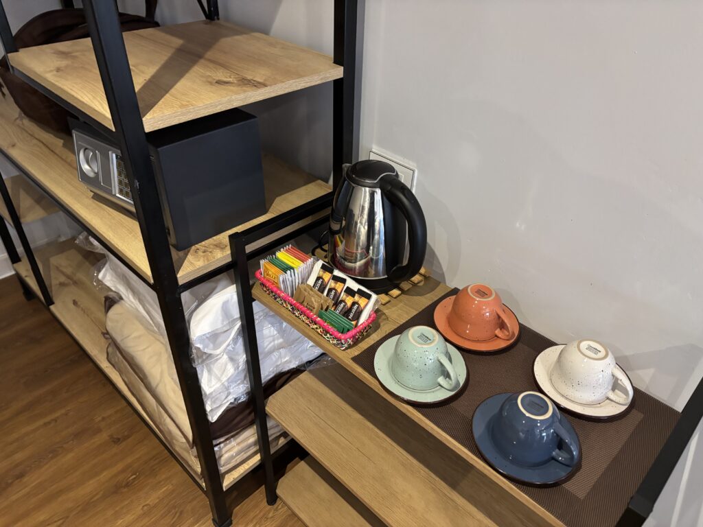 The image shows a wooden shelf unit with several items. On the top shelf, there is a black safe. Below it, there are folded blankets or bedding. On the right side of the image, there is a wooden surface with a stainless steel electric kettle, a colorful basket containing various tea and coffee packets, and four ceramic cups with matching saucers in different colors: orange, light blue, white, and dark blue. The surface is covered with a brown mat.