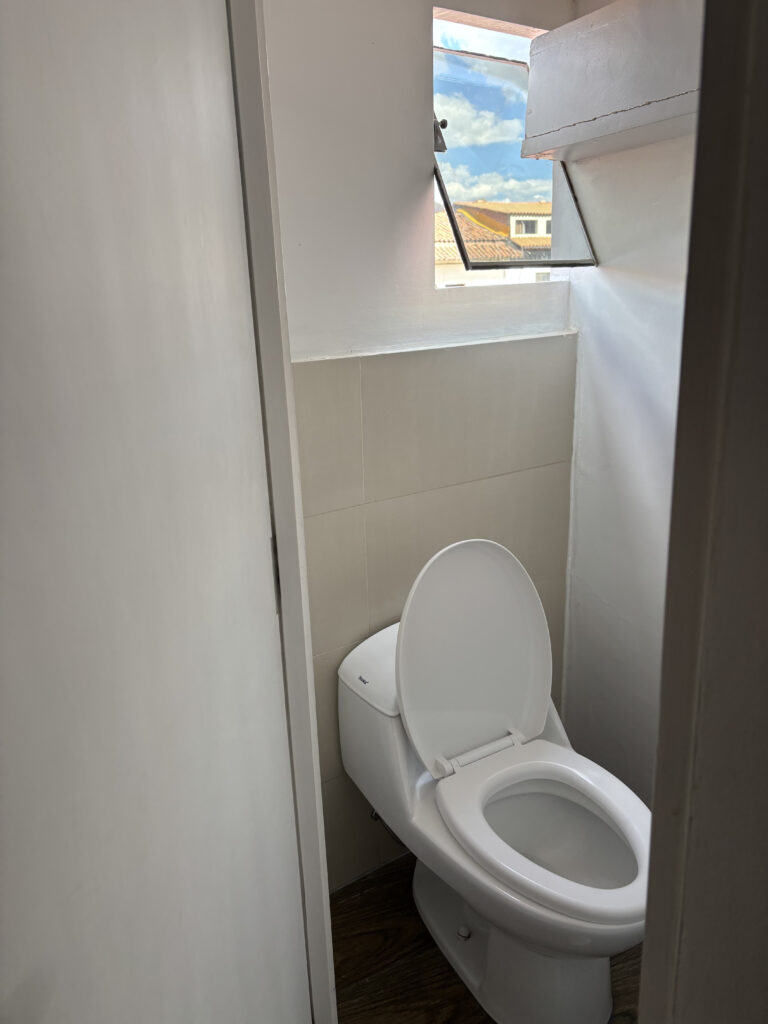 The image shows a small bathroom with a white toilet. The toilet seat is open, and there is a window above it, allowing natural light to enter. The window is slightly open, and the view outside shows a blue sky with clouds and part of a building with a tiled roof. The walls and door are white, and the floor appears to be wooden.