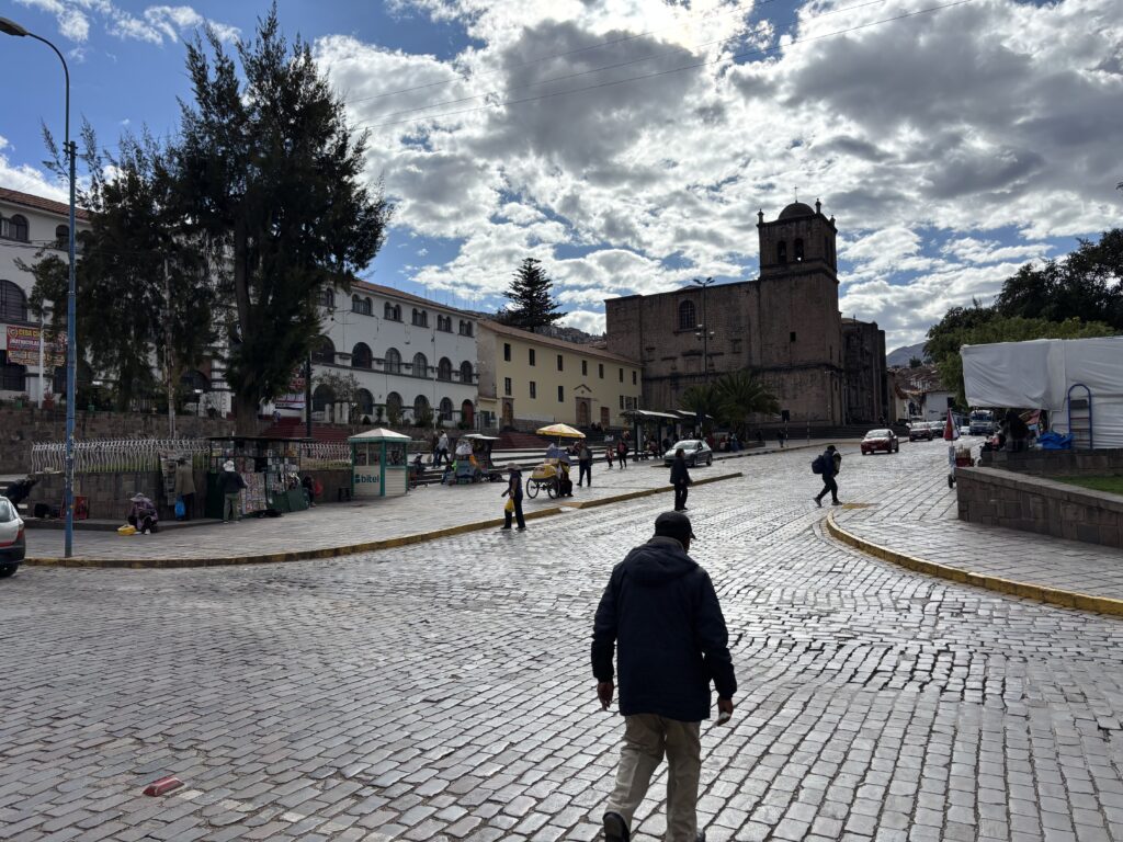 A cobblestone street in a historic area with people walking and a few cars. On the left, there are trees and a white building with arched windows. In the background, a large stone church with a bell tower is visible. The sky is partly cloudy, and there are small vendor stalls along the street.