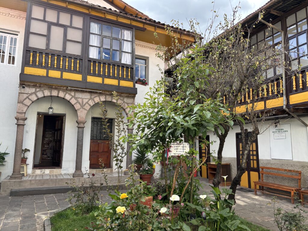 The image shows a charming courtyard with a traditional building featuring wooden balconies and yellow accents. The building has arched doorways and windows with decorative railings. In the foreground, there is a garden with various plants and flowers, including roses and calla lilies. A sign is visible among the plants, and a wooden bench is placed against the building. The sky is partly cloudy.