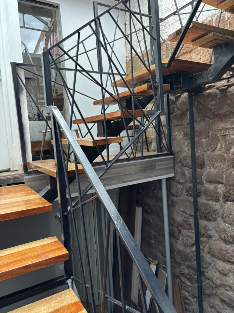 The image shows a modern staircase with wooden steps and a black metal railing featuring a geometric design. The staircase is situated indoors, adjacent to a stone wall. There is a glass door at the top of the stairs, leading to another area. The overall setting appears to be a mix of industrial and rustic styles.