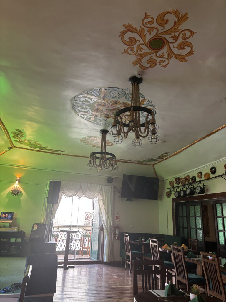 The image shows the interior of a restaurant or dining area with wooden tables and chairs. The ceiling is decorated with ornate patterns and two chandeliers with exposed bulbs. There is a large window with sheer curtains, allowing natural light to enter. A television is mounted on the wall, and various decorative masks are displayed above a doorway. The floor is wooden, and there is a small stage area with audio equipment on the left side.