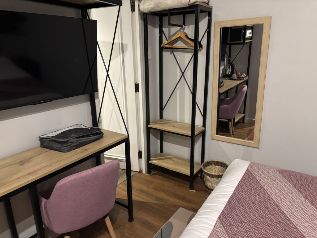 The image shows a neatly arranged room with a modern design. There is a wooden desk with a black metal frame, a flat-screen TV mounted above it, and a pink chair in front. On the desk, there is a gray bag. To the right, there is an open wardrobe with shelves and a few wooden hangers. A full-length mirror with a wooden frame is mounted on the wall next to the wardrobe. A wicker basket is placed on the floor near the wardrobe. Part of a bed with a patterned pink and white bedspread is visible in the foreground. The room has wooden flooring.