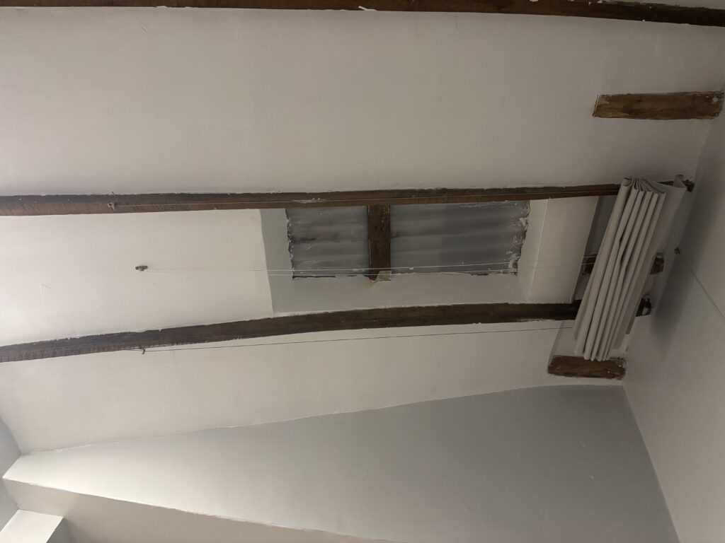 The image shows a ceiling with exposed wooden beams. There is a section with a translucent panel, possibly a skylight or a covered opening. A folded curtain or blind is attached to one side of the ceiling. The walls and ceiling are painted white.