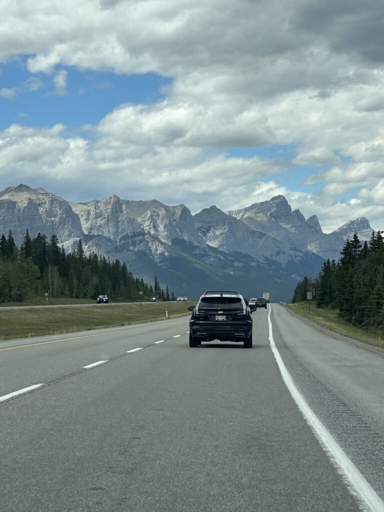 A scenic highway view with a black SUV driving on a road surrounded by lush green trees on both sides. In the background, there are majestic, rugged mountains under a partly cloudy sky with patches of blue.
