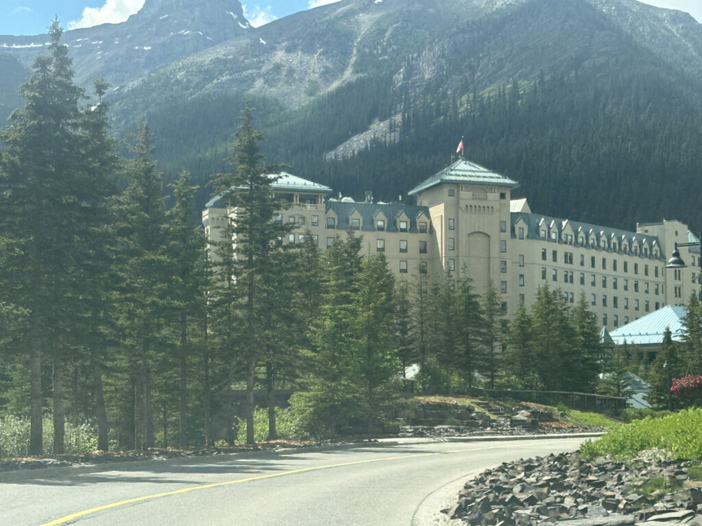A large, elegant hotel building with a light-colored facade and multiple windows is partially obscured by tall evergreen trees. The hotel has a distinctive roof with a flag on top. In the background, there are towering mountains with patches of snow and dense forest. A winding road leads towards the hotel, bordered by rocks and greenery. The sky is clear with a few clouds.