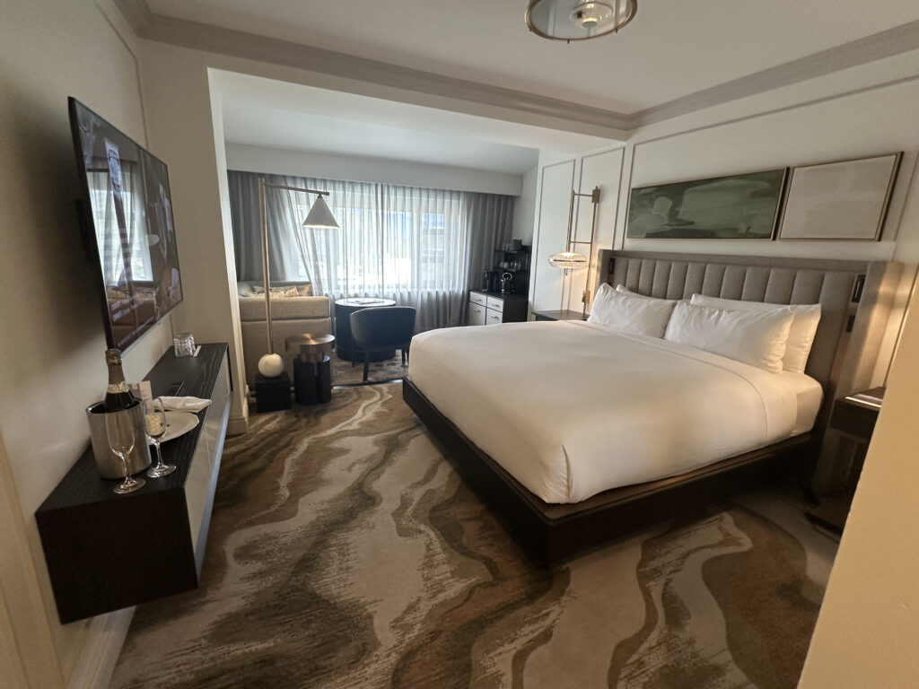 A modern hotel room with a large bed featuring white linens and a padded headboard. On the left, a wall-mounted TV is above a console with a champagne bottle, glass, and plate. The room has a patterned carpet and a seating area by the window with a chair, small table, and floor lamp. The window is covered with sheer curtains, and there is artwork above the bed.