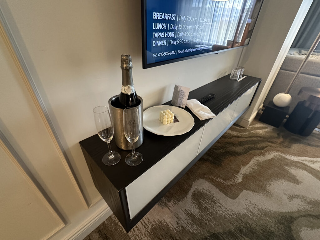 The image shows a hotel room setup with a dark wooden console table. On the table, there is a bottle of champagne in a metal ice bucket, two empty champagne flutes, and a plate with a small serving of butter or cheese. A folded napkin and a small card are also on the table. A television is mounted on the wall above the console, displaying dining information. The room has a patterned carpet and modern decor.
