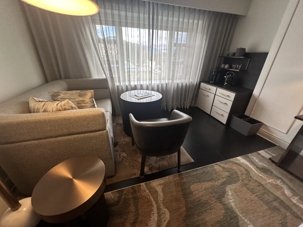 The image shows a cozy hotel room corner with a light gray L-shaped sofa adorned with beige pillows. In front of the sofa is a round coffee table and a single armchair. A large window with sheer curtains allows natural light to fill the room. To the right, there is a small kitchenette area with a coffee maker, cups, and a set of drawers. A modern lamp hangs above the seating area, and a patterned carpet covers the floor.