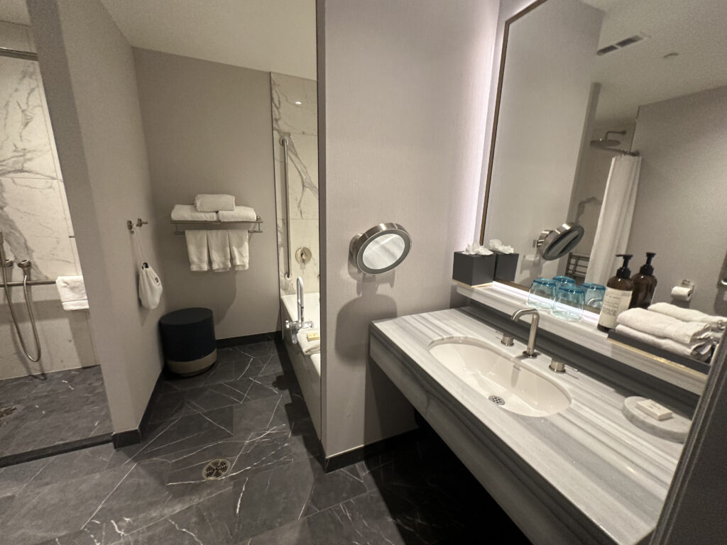 The image shows a modern bathroom with a marble floor and walls. On the right, there is a sink with a large mirror above it, accompanied by a magnifying mirror. The countertop holds two blue glasses, soap, and bottles of toiletries. On the left, there is a shower area with a handheld showerhead and a bathtub with a towel rack above it, holding neatly folded white towels. The overall design is sleek and contemporary.