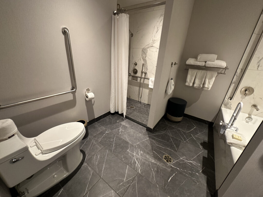 The image shows a modern bathroom with gray marble flooring. On the left, there is a toilet with a safety grab bar next to it and a roll of toilet paper mounted on the wall. In the center, there is a shower area with a curtain and a handheld showerhead, also equipped with grab bars. On the right, there is a sink with a mirror above it, and a shelf holding neatly folded white towels. A small trash bin is placed on the floor near the shower.