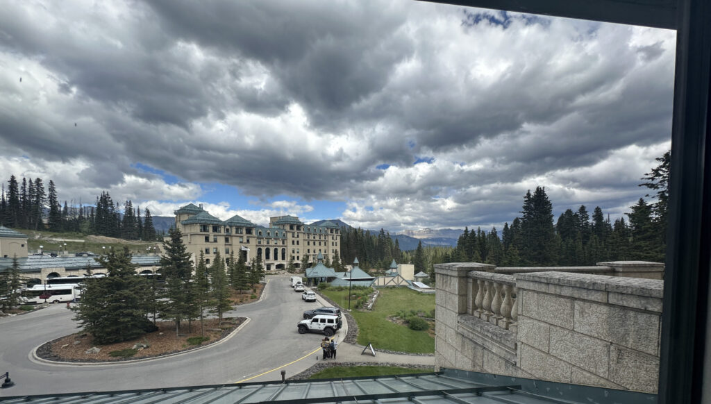 A scenic view from a window showing a large, elegant building with multiple stories and a green roof, surrounded by a circular driveway. Several vehicles, including a white van and a white SUV, are parked along the driveway. The area is landscaped with tall evergreen trees and a grassy lawn. In the background, there are more trees and distant mountains under a cloudy sky. The foreground includes part of a stone balcony and a metal roof.
