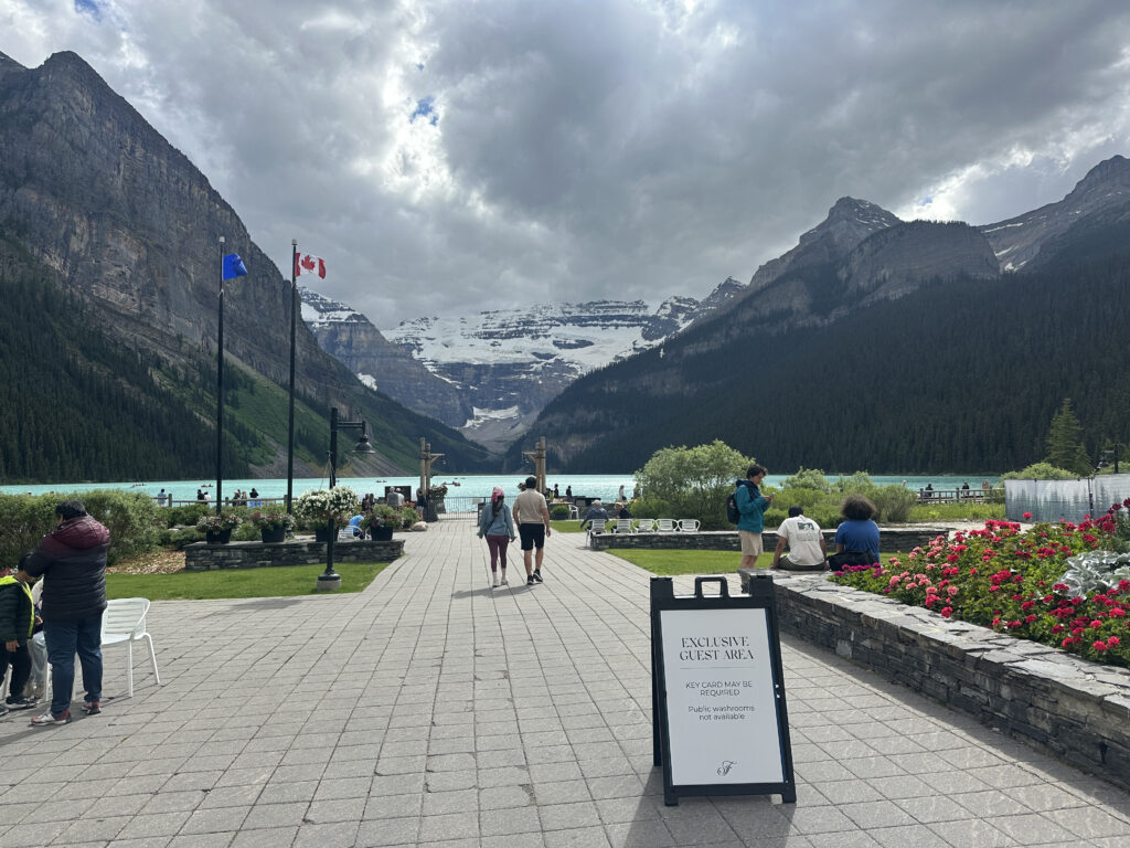 The image shows a scenic view of a lake surrounded by mountains, with a cloudy sky above. In the foreground, there is a paved walkway with people walking and sitting. A sign reads "Exclusive Guest Area" with additional text about key card access and public washrooms. There are two flags, one of which is the Canadian flag, and flower beds with red and pink flowers. The lake has a turquoise color, and the mountains in the background are partially covered with snow.