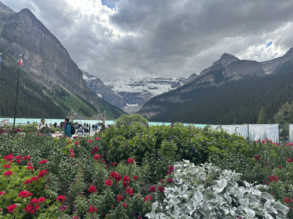 A scenic view of a mountainous landscape with a turquoise lake in the background. The foreground features a vibrant garden with red and pink flowers and lush green foliage. People are visible near the lake, and the sky is overcast with dramatic clouds. Snow-capped peaks and dense forests surround the lake, creating a picturesque setting.