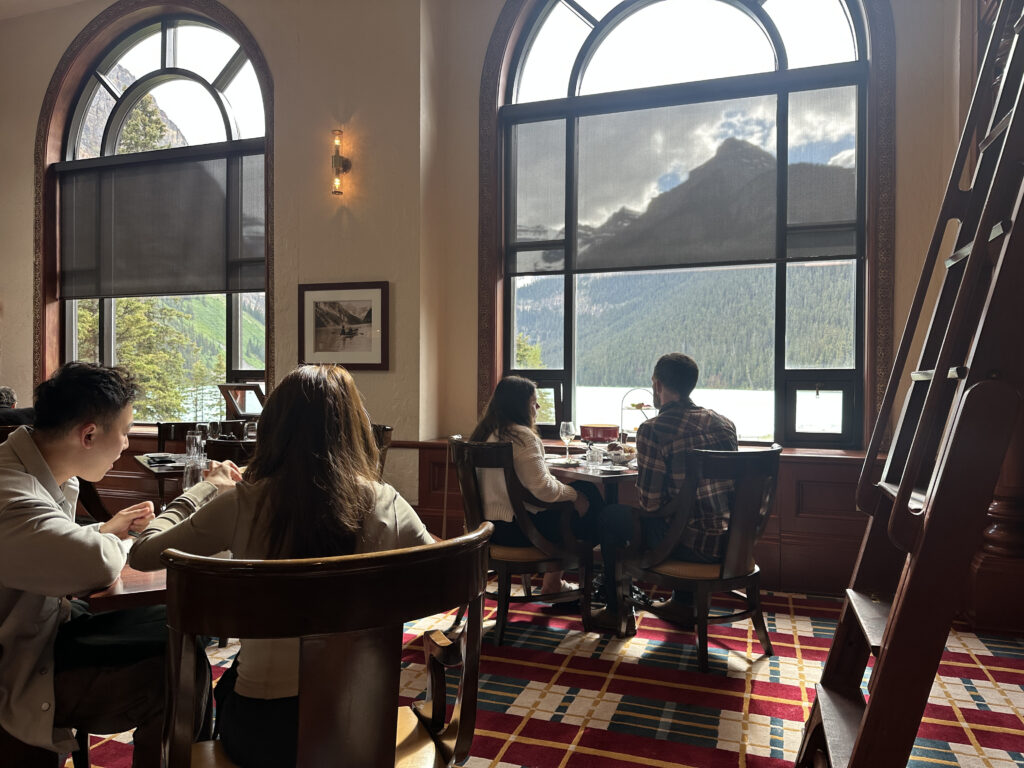 A group of people is seated at a table in a restaurant with large arched windows. The windows offer a view of a scenic landscape featuring a lake and mountains. The interior has a warm, cozy atmosphere with a patterned carpet and wooden furniture. A ladder is visible on the right side of the image.