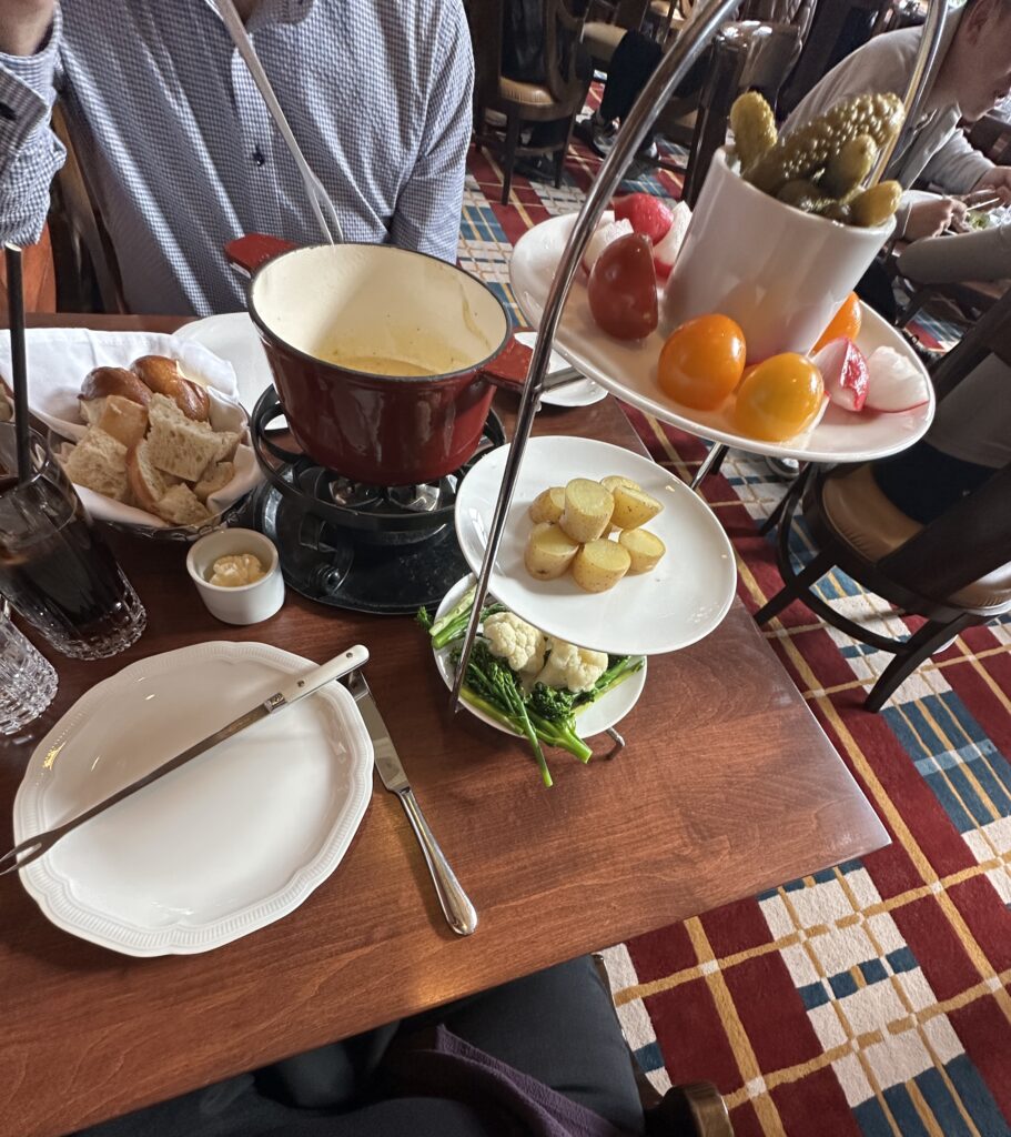 A table setting with a fondue pot containing melted cheese, surrounded by various foods for dipping. There is a plate of bread pieces, a small dish of sauce, and a glass of dark soda. A tiered stand holds pickles, cherry tomatoes, radishes, boiled potatoes, and a bowl of vegetables including cauliflower and broccolini. A person in a checkered shirt is partially visible, sitting at the table.