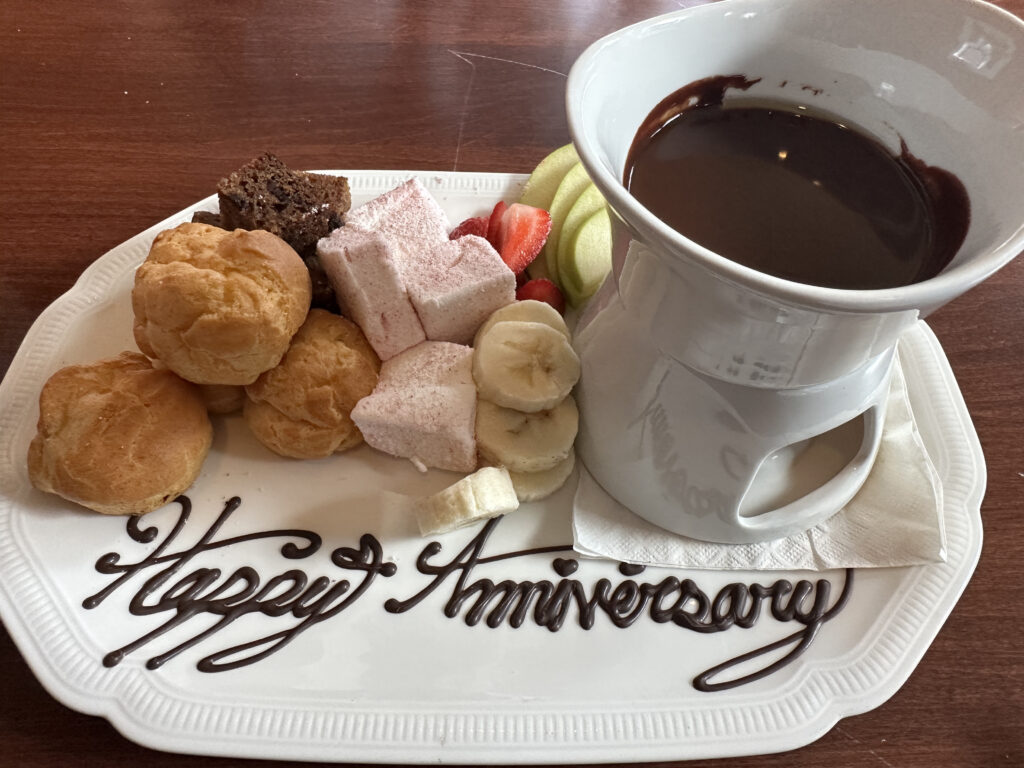 A white plate with "Happy Anniversary" written in chocolate sauce. The plate contains a variety of treats, including cream puffs, pink marshmallows, banana slices, strawberries, apple slices, and a piece of chocolate cake. There is also a white fondue pot filled with chocolate sauce on the plate.