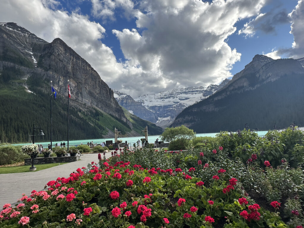 A scenic view of a mountainous landscape with a lake in the background. The foreground features vibrant red and pink flowers in a garden. Two flags are visible near a pathway, and people are gathered near the lake. The sky is partly cloudy, with sunlight illuminating the scene.