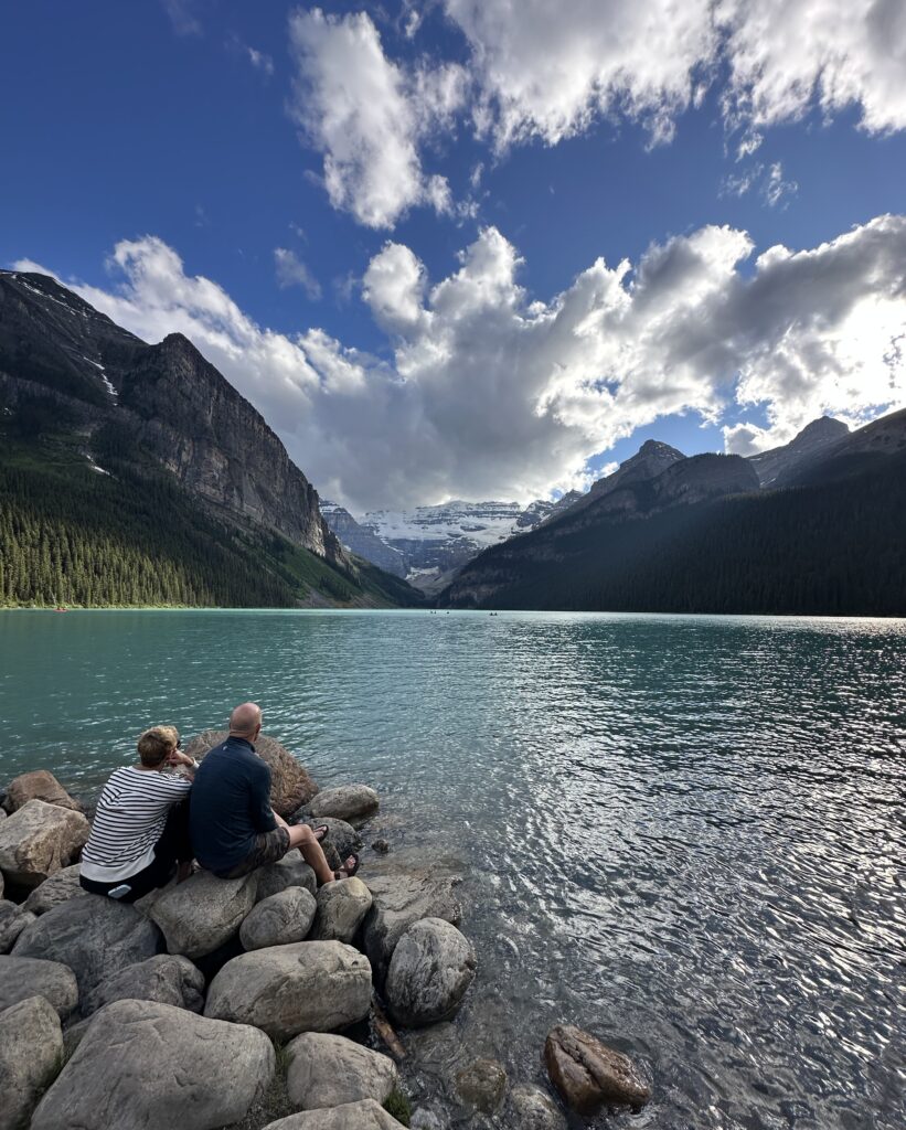 A couple is sitting on rocks by a turquoise lake, surrounded by mountains. The sky is partly cloudy, and the scene is serene and picturesque.