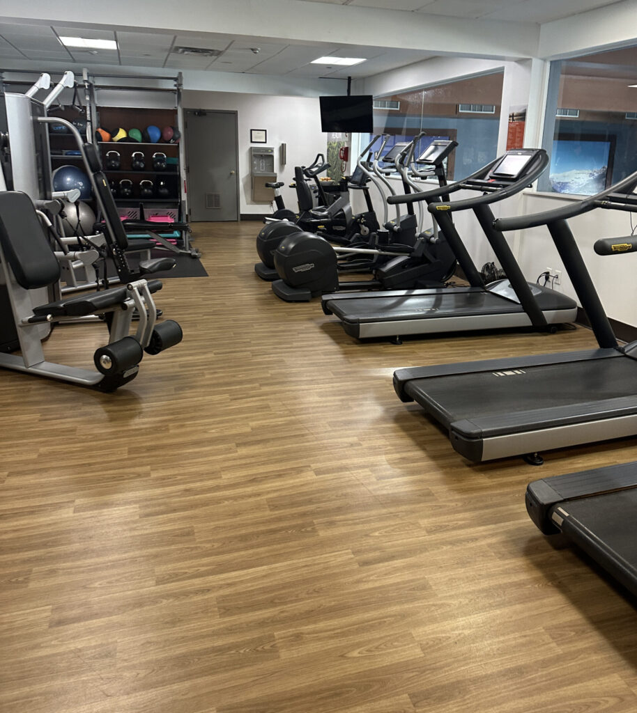 The image shows a gym with various exercise equipment. On the right, there are several treadmills lined up against a window. In the center, there are stationary bikes. On the left, there is a weight machine and a rack with colorful exercise balls and weights. The floor is wooden, and the ceiling has recessed lighting.