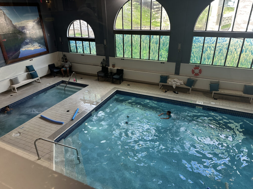 The image shows an indoor swimming pool area with two pools. One is a larger swimming pool where a person is swimming, and the other is a smaller hot tub with a person sitting in it. The area is surrounded by large windows with scenic views of greenery and mountains. There are benches with cushions along the walls, and a large landscape painting is visible on one wall. A pool noodle is lying on the edge of the hot tub.